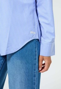 Chemise à manches longues rayée bleu clair avec un ourlet arrondi et un logo brodé sur le côté, assortie à un jean bleu moyen.