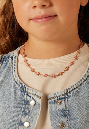 Goldene Kettenhalskette mit kleinen, pinken Blumenanhängern. getragen über einem cremefarbenen Pullover, ergänzt durch eine Jeansjacke.