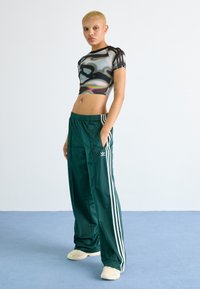 Graphic cropped shirt with wavy gray-black pattern, paired with dark green track pants featuring white side stripes and logo, on a plain background.