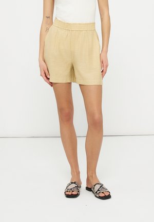 Beżowe szorty z elastycznym pasem noszone z białym topem i czarnymi, otwartymi klapkami w wzór skóry węża na stojącej osobie na białym tle.