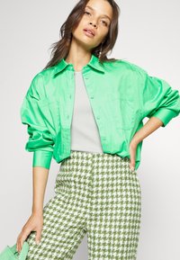Camisa oversized de cor verde brilhante, com dois bolsos, usada sobre uma blusa cinza e calças com padrão houndstooth verde e branco.