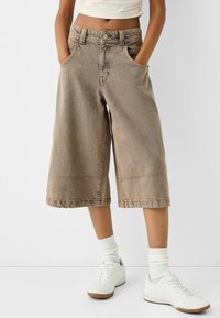 Culottes en denim marron avec une coupe loose, taille mi-haute et ourlet effiloché. Associés à des baskets blanches et des chaussettes montantes.