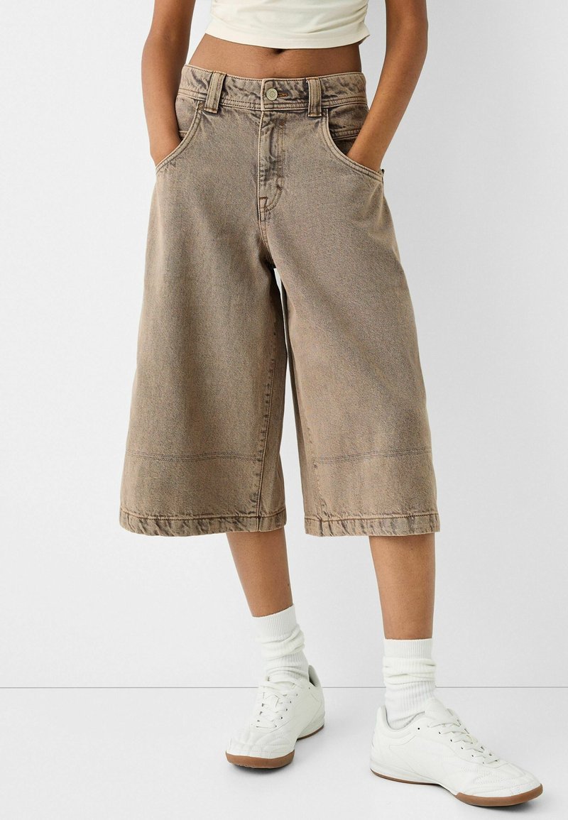 Culottes en denim marron avec une coupe loose, taille mi-haute et ourlet effiloché. Associés à des baskets blanches et des chaussettes montantes.