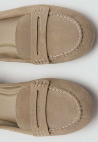 Mocasines de ante beige con un diseño cosido y una correa decorativa. Suela plana, punta redondeada, textura suave.