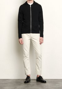 Svart zip-up hoodie med en framficka, parat med vita slim-fit byxor och svarta dressade skor på ett grått golv.