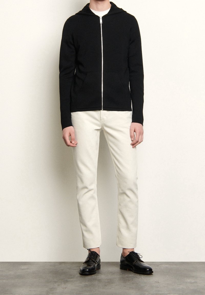 Svart zip-up hoodie med en framficka, parat med vita slim-fit byxor och svarta dressade skor på ett grått golv.