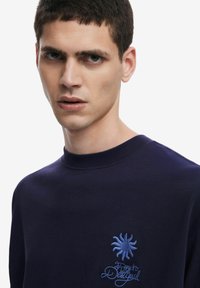Sudadera azul marino de algodón, con cuello redondo, diseño de sol bordado y texto en azul claro en la parte izquierda del pecho.
