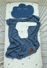 Blaues gewebtes Wickel mit einem weißen Langarm-Overall mit grauen Sternenprint, über einer sternenmusternden Matratze des Kinderbettes und einem wolkenförmigen Kissen.