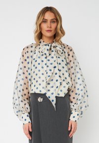 Blusa trasparente con scollatura a fiocco, caratterizzata da un motivo a pois in diverse tonalità di blu su uno sfondo crema; maniche lunghe a sbuffo.