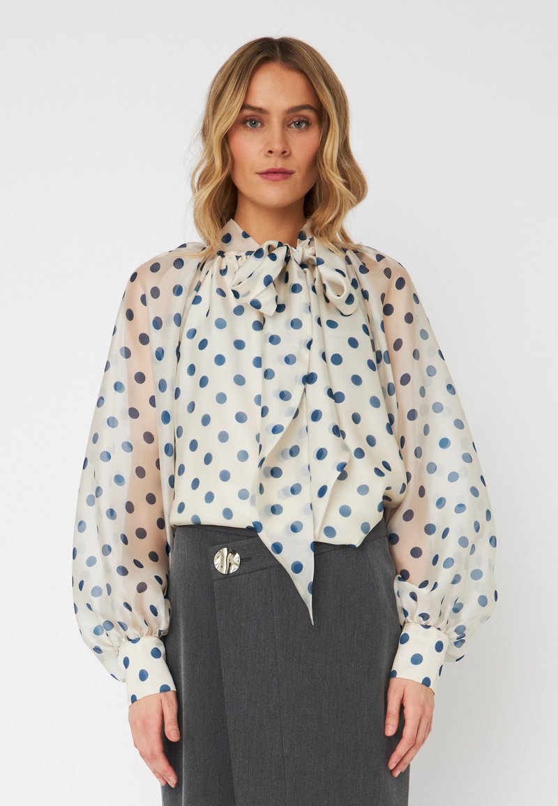 Blusa trasparente con scollatura a fiocco, caratterizzata da un motivo a pois in diverse tonalità di blu su uno sfondo crema; maniche lunghe a sbuffo.