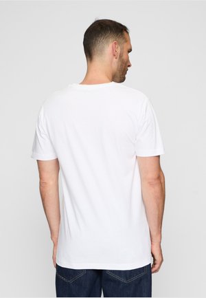 Muž oblečený v jednoduchom bielom tričku s krátkym rukávom a tmavomodrých džínsach, zobrazený zozadu na jednoliatom svetlosivom pozadí.