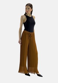 Bruine geplooide broek met wijd uitlopende pijpen, een elastische tailleband en trekkoord, gecombineerd met een aansluitend zwart mouwloos topje en puntige zwarte schoenen.