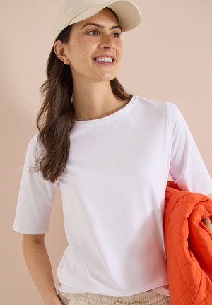 Femme aux longs cheveux bruns portant une chemise blanche et une casquette beige, tenant une veste matelassée orange sur son bras, souriante, fond beige.