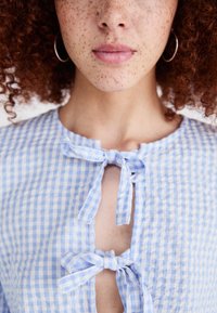 Μπλέ και λευκό καρό μπλούζα κατασκευασμένη από ελαφρύ ύφασμα, με κλείσιμο με κορδόνια στον λαιμό και ανοιχτό σχέδιο κλειδώματος.