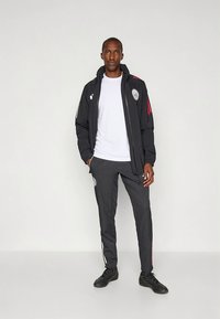 Svart sportjacka med röda detaljer och logotyp, kombinerad med grå byxor och svart skor, lämplig för sport eller vardagsbruk.