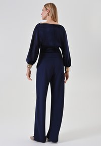 Vrouw die met haar rug naar de camera staat, gekleed in marineblauwe wijdvallende broek en een bijpassend blouse met lange mouwen, vergezeld van gouden armbanden en oorbellen.