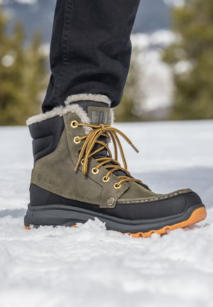 Botas de invierno verdes y negras con forro de pelaje sintético, ojales dorados, cordones negros y suela naranja. Presenta una parte superior texturizada y un talón reforzado.