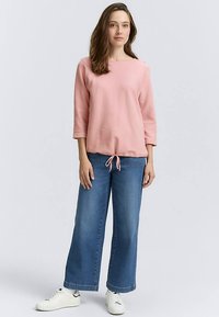 Roze top met lange mouwen en een ronde halslijn met trekkoord aan de zoom, gecombineerd met wijd uitlopende blauwe spijkerbroek en witte sneakers.