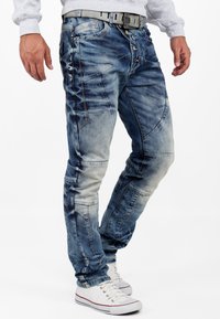 Homme portant un jean bleu décoloré et usé avec fermeture à boutons, une ceinture grise, un sweat-shirt gris et des baskets blanches, se tenant sur un fond blanc.