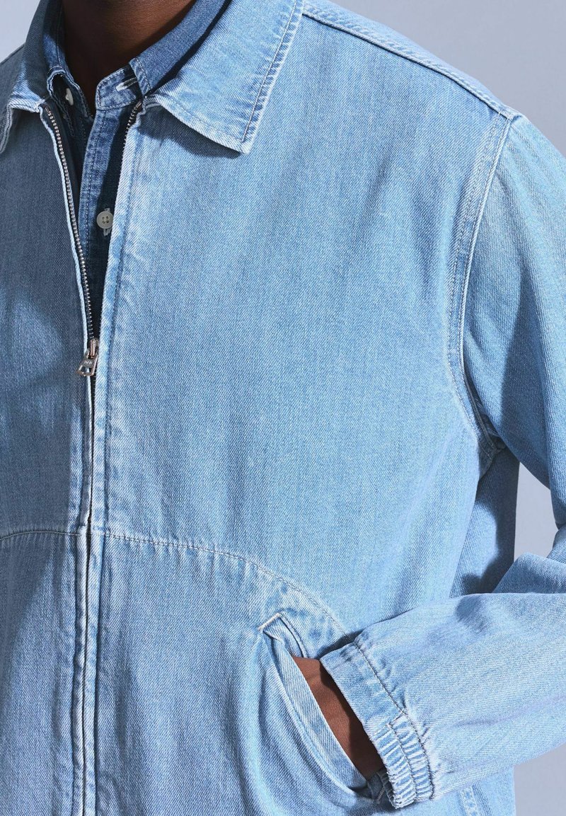 Hellblaue Denim-Jacke mit Reißverschluss vorne, spitzem Kragen und seitlichen Taschen. Sie hat sichtbare Nähte und eine lockere Passform.