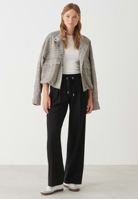 Grijze geblokte cropped jas met knoopdetails, witte nauwsluitende blouse en zwarte wijdvallende broek; gecombineerd met witte sneakers.