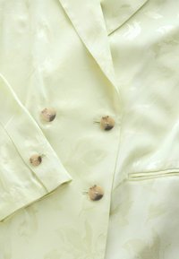 Blazer crème clair à double boutonnage avec un motif floral subtil, quatre boutons marron et une poche fendue sur le côté droit.