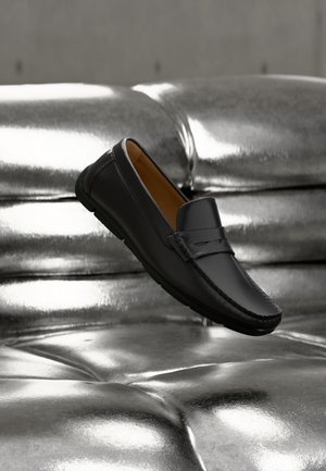Schwarzer Lederloafer, der über einem glänzenden silberfarbenen, gesteppte Ledersofa mit weicher Beleuchtung schwebt.