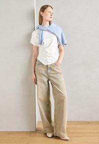 Hvit t-skjorte med korte ermer, løse beige jeans med lyseblå sømmer, lyseblå genser drapert rundt skuldrene, kremfargede sko, tregulv.