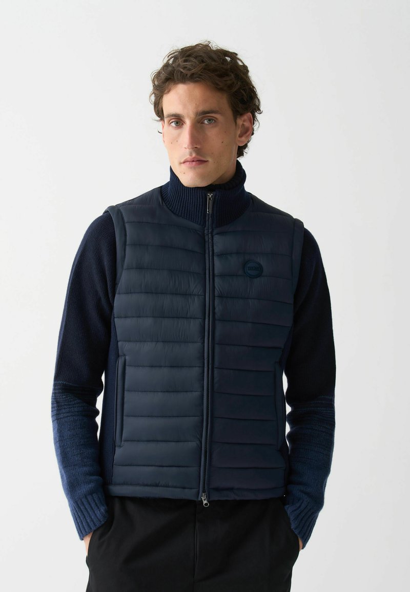 Námornícky puffer vest s horizontálne prešívanými časťami, vysokým golierom, plným predným zipsom a detailom loga. Nosí sa na tmavom svetri s textúrovanými rukávmi.