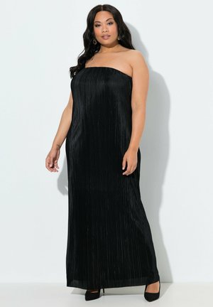 Frau mit langen dunklen Haaren, die ein trägerloses, schwarzes, plissiertes bodenlanges Kleid und schwarze High Heels trägt, steht vor einer weißen Wand.