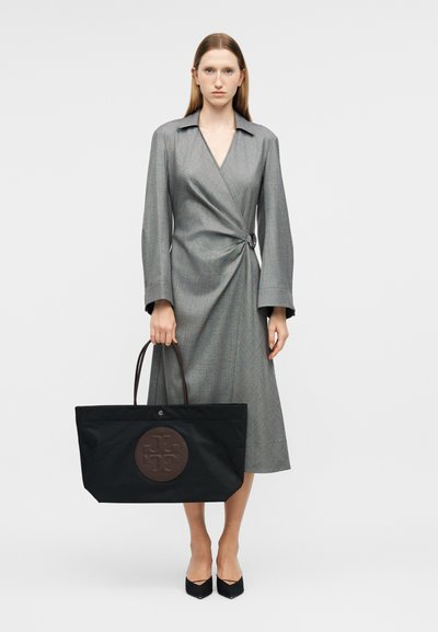 Femme portant une robe cache-cœur grise tenant un grand sac cabas noir avec un logo circulaire brun foncé, debout devant un fond blanc.
