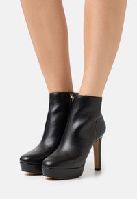 Botines de cuero negro con puntera redondeada, tacón de aguja alto, plataforma delantera y detalle de cremallera lateral. Textura lisa y diseño elegante.