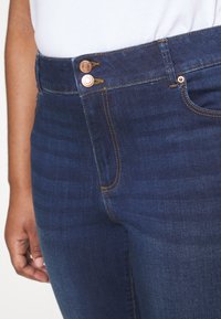 Høytlivs denim jeans i mørkeblå, med to kobberknapper, kontrasterende gul søm og en jevn tekstur.