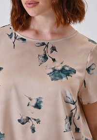 Frau mit beiger Bluse mit kurzen Ärmeln und blau-grauem Blumenmuster, von den Schultern bis zur oberen Brust gezeigt.
