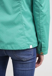 Tyrkysová bunda s malými bílými puntíky nošená přes modré džínové kalhoty, ukazující záda v pase a pravou ruku odpočívající vedle boků.