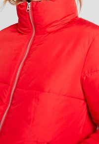 Red puffer jacket with a high collar, featuring a front zipper and stitched sections for insulation. Smooth, shiny fabric texture.