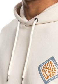 Béžová mikina s kapucí a nastavovacími šňůrkami, s dvoubarevným logem Quiksilver v oranžové a modré barvě. Měkký bavlněný materiál, volný design.