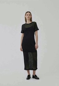 Vestido preto de renda com mangas curtas, apresentando padrões geométricos, uma silhueta ajustada e uma bainha mais comprida, combinado com sapatos pretos.