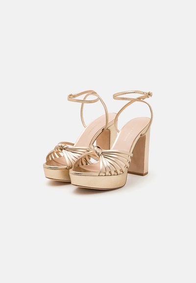 Sandalias doradas de tacón alto con plataforma, con un diseño de tiras entrelazadas, correas ajustables en el tobillo y un detalle de nudo en la parte delantera.