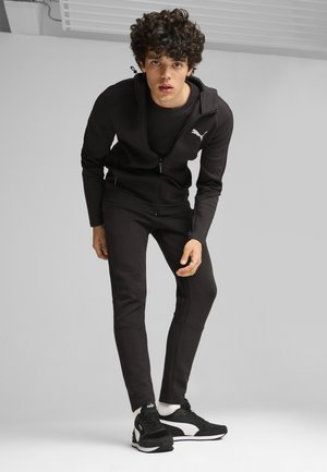 Junger Mann, der sich nach vorne beugt, trägt einen schwarzen Puma-Trainingsanzug mit Sneakers vor einem schlichten hellen Hintergrund.