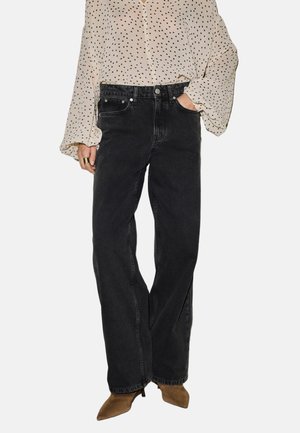 Osoba nosiaca voľné čierne džínsy, béžové špicaté čižmy a krémovú blúzku s čiernymi bodkami, stojaca s rukou v vrecku.