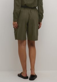 Personne portant une chemise à manches longues vert olive et un short assorti arrivant aux genoux, avec des sandales noires à glissement, se tenant devant un fond blanc uni.