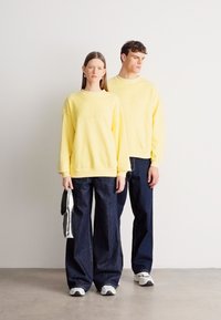 Κίτρινο φούτερ με ανάγλυφα γράμματα, σε συνδυασμό με χαλαρό σκούρο τζιν παντελόνι. Το μοντέλο στα αριστερά κρατά μια τσάντα· και οι δύο φορούν αθλητικά παπούτσια.