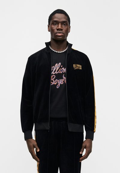 Μαύρο βελούδινο μπουφάν με φερμουάρ, με χρυσές λεπτομέρειες και λογότυπο, συνδυασμένο με μαύρο μπλουζάκι. Το μοντέλο φοράει ταιριαστό μαύρο παντελόνι.