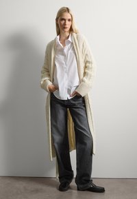 Larga chaqueta de punto trenzado en crema, superpuesta sobre una camisa blanca de botones. Combinada con jeans oscuros de corte holgado y zapatos negros.