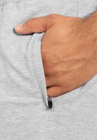 Sudadera gris con un bolsillo en el que hay una mano colocada dentro. El bolsillo tiene una cremallera visible con herrajes negros. La textura de la tela parece suave y lisa.