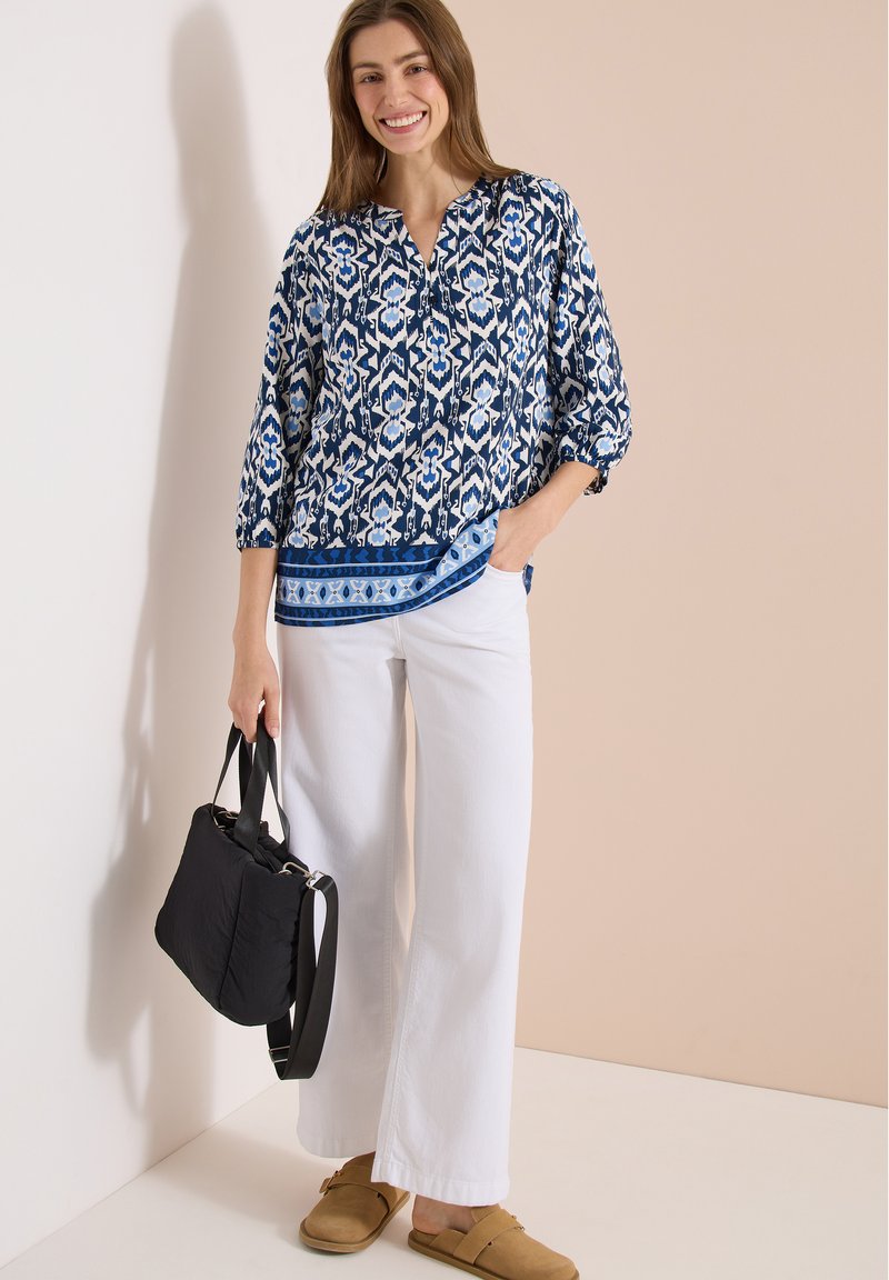 Jeune femme debout souriante portant un chemisier bleu à motifs, un pantalon blanc à jambes larges, des chaussures marron slip-on et tenant un sac à main noir.