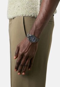 Srebrni metalni sat s okruglim plavim brojčanikom, koji ima tri podbrojčanika, nosi se na zapešću ispred pozadine svijetlozelenog pletenog pulovera i smešnih hlača.