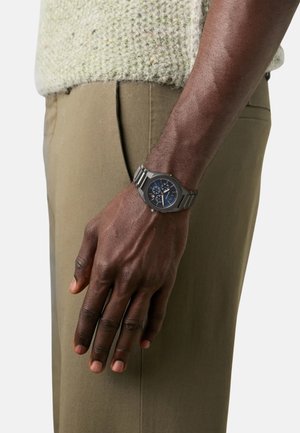 Montre en métal argenté avec un cadran bleu rond, dotée de trois sous-cadrans, portée au poignet sur un fond de pull en tricot vert clair et de pantalons beiges.