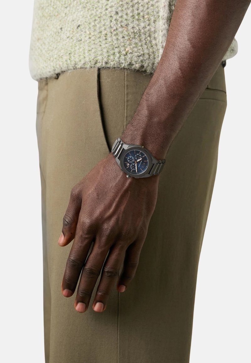 Srebrni metalni sat s okruglim plavim brojčanikom, koji ima tri podbrojčanika, nosi se na zapešću ispred pozadine svijetlozelenog pletenog pulovera i smešnih hlača.